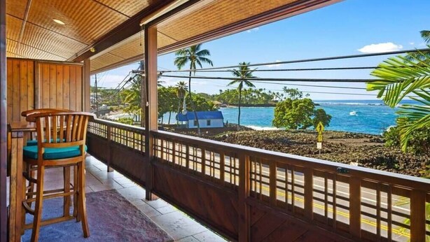 Un balcón con una silla de madera y vista al océano y palmeras.