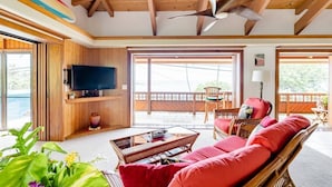 Amplia sala de estar con televisor grande, vigas de madera en el techo, ventilador de techo y vistas al exterior.