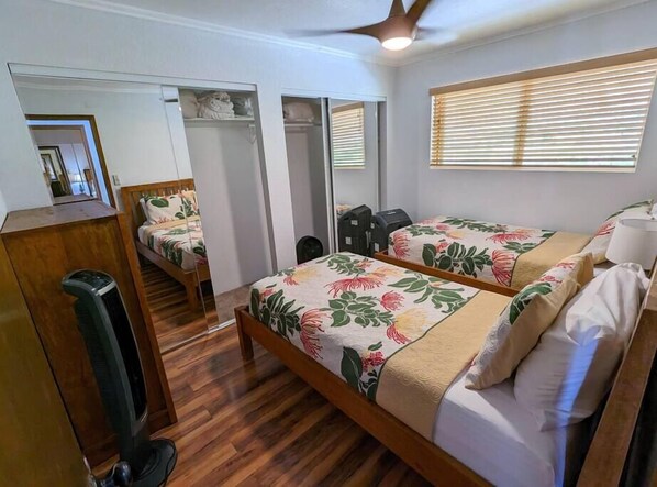 A bedroom with two beds, wooden flooring, a ceiling fan, and a mirror. - Sunset Oceanside Condo (Kahaluu-Keauhou)