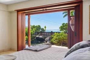 Een slaapkamer met een groot schuifglasraam dat uitkomt op een balkon met uitzicht op de oceaan en tropische begroeiing.
