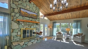 A spacious living room with a stone fireplace, wooden ceiling beams, and large windows offering a view of the outdoors.