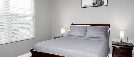 Ein Schlafzimmer mit einem Bett, zwei Nachttischen, einem Fenster mit Jalousien und einem gerahmten Bild an der Wand.