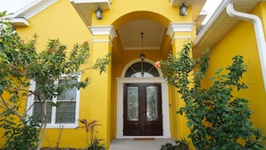 Una casa amarilla con puerta marrón oscuro, detalles blancos y un pequeño porche con plantas.