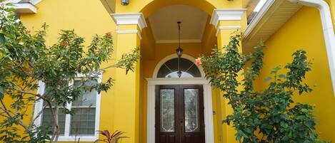 A yellow house with a dark brown door, white trim, and a small porch area with plants.