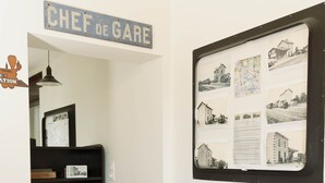A room with a wooden chair, a desk with a computer, a wall-mounted sign reading "CHEF DE GARE", and a bulletin board with photos and text.