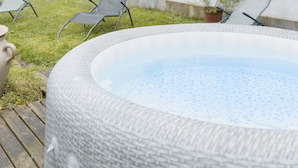 An outdoor hot tub area with two lounge chairs, a wooden deck, and a white building with red shutters in the background.