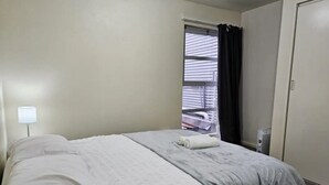Una cama bien hecha con ropa blanca y una toalla enrollada, una ventana con persianas, y una puerta a la derecha.