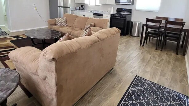 Ein geräumiger Wohnbereich mit einer großen, beigen Couch, einem runden Couchtisch und einem Esstisch mit Stühlen. Im Hintergrund ist eine Küche mit weißen Schränken und Geräten erkennbar.