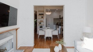 Un salon moderne avec un mur en briques blanches, un coin repas avec une table ronde et des chaises, et une cuisine en arrière-plan.