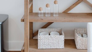 Une étagère en bois sur laquelle sont disposés divers objets, notamment un bol contenant des pommes vertes, un mortier et un pilon, ainsi que des bocaux en verre fermés par des bouchons en liège.