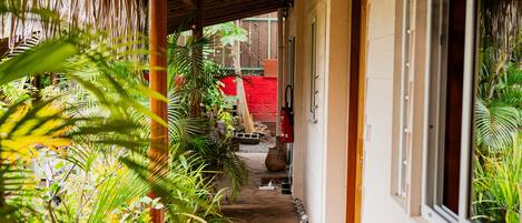 A covered walkway with stone flooring, surrounded by lush greenery and a wooden ceiling structure.