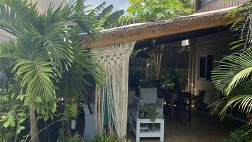 Un patio couvert avec un canapé en osier blanc, des plantes en pot et un chemin en pierre.