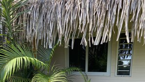 Uma casa com telhado de palha, rodeada por vegetação exuberante, incluindo palmeiras e plantas floridas.