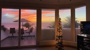 Een kamer met grote ramen met uitzicht op de ondergaande zon boven de oceaan, een kerstboom en een televisie op een standaard.