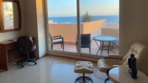 Een kamer met een bureau, een bureaustoel en een bank. Er is een grote schuifdeur die leidt naar een buitenbalkon met uitzicht op de oceaan.
