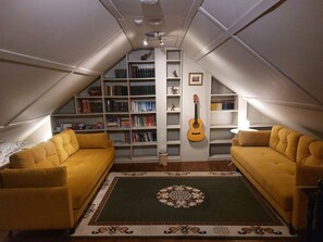 Une pièce mansardée chaleureuse, avec un canapé jaune, un tapis à motifs et une bibliothèque remplie de livres et d’une guitare.