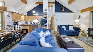 A spacious living area with a large blue sofa, a wooden bench, and a dining table set with plates and glasses. A kitchen with wooden cabinets and a dining area with a blue couch and a fireplace are visible in the background.