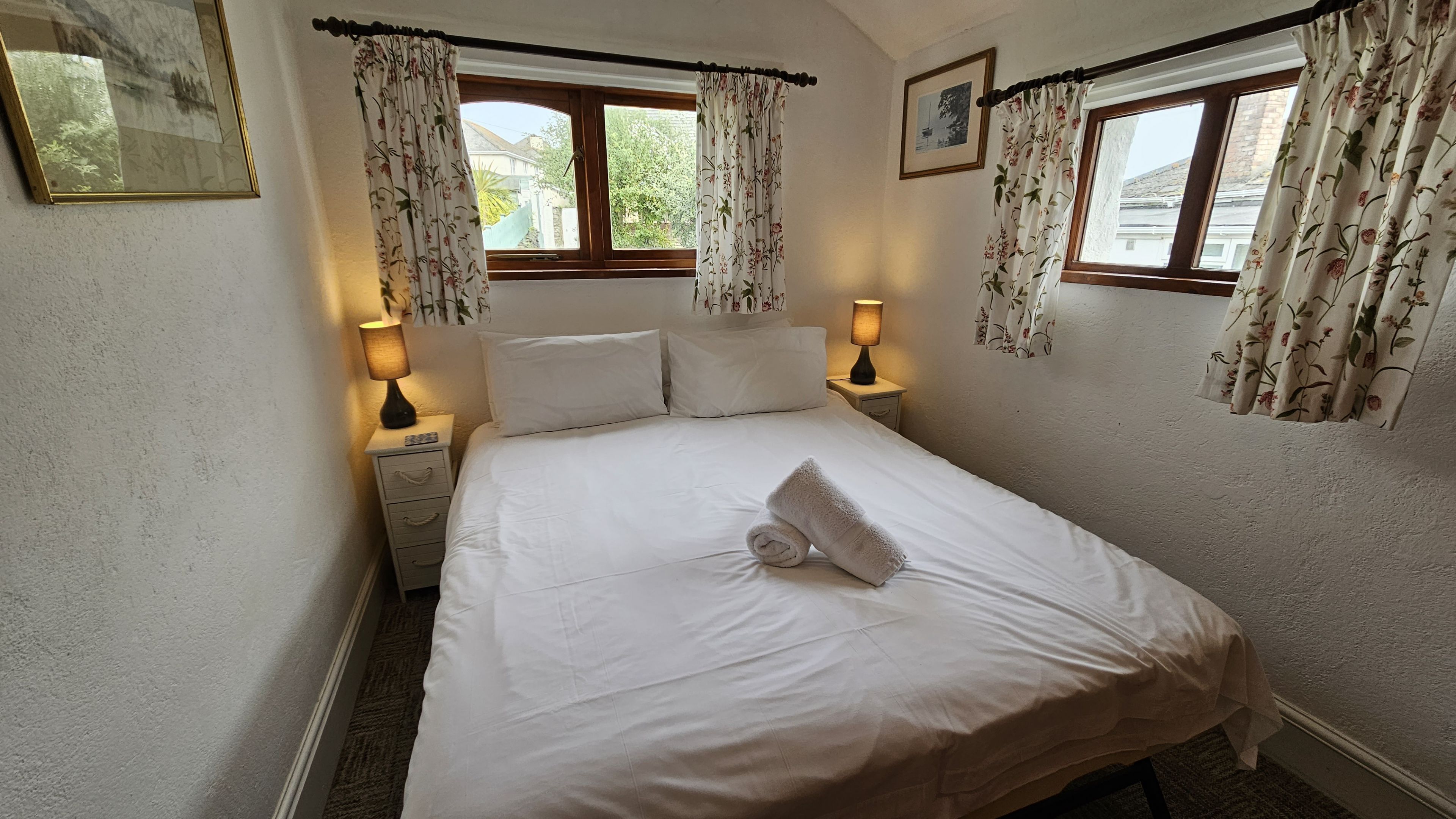 Een net opgemaakt bed met een opgerolde handdoek, twee bedlampjes en bloemen gordijnen voor een raam.