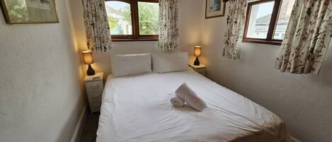 Een net opgemaakt bed met een opgerolde handdoek, twee bedlampjes en bloemen gordijnen voor een raam.