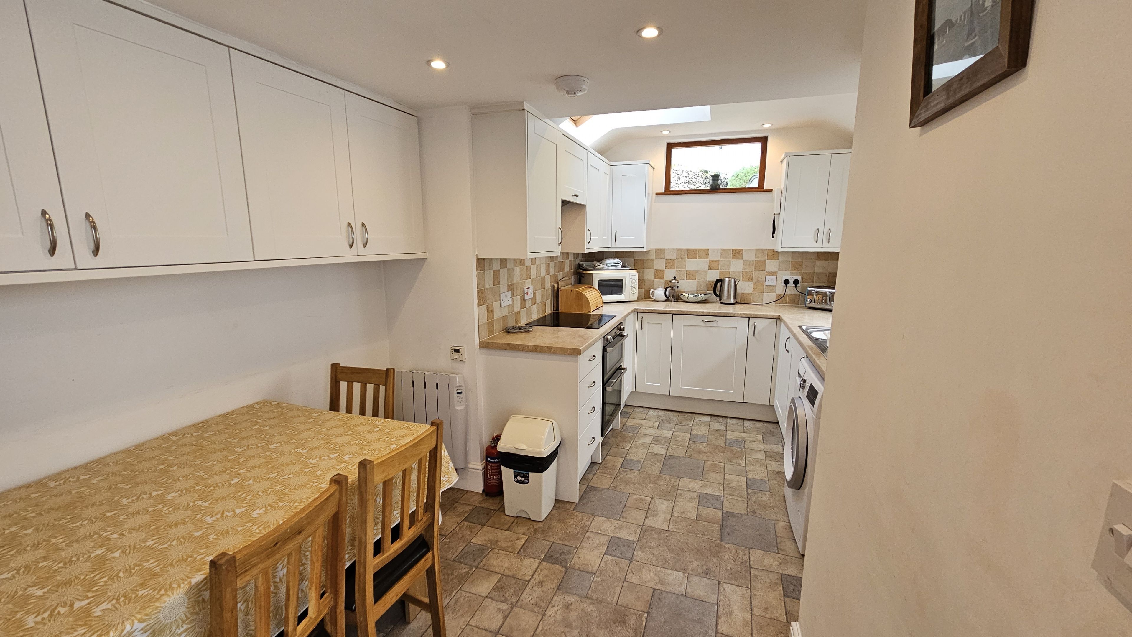 A compact kitchen with white cabinets, a wooden dining table, and a small trash bin.