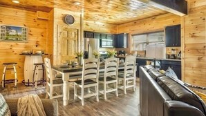A cozy cabin interior with a wooden interior, a dining area with white chairs, a black leather couch, and a kitchen with dark cabinets.