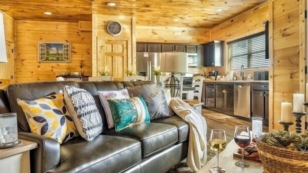 A cozy cabin interior with a leather sofa, wooden walls, a dining area with a table and candles, and a kitchen in the background.