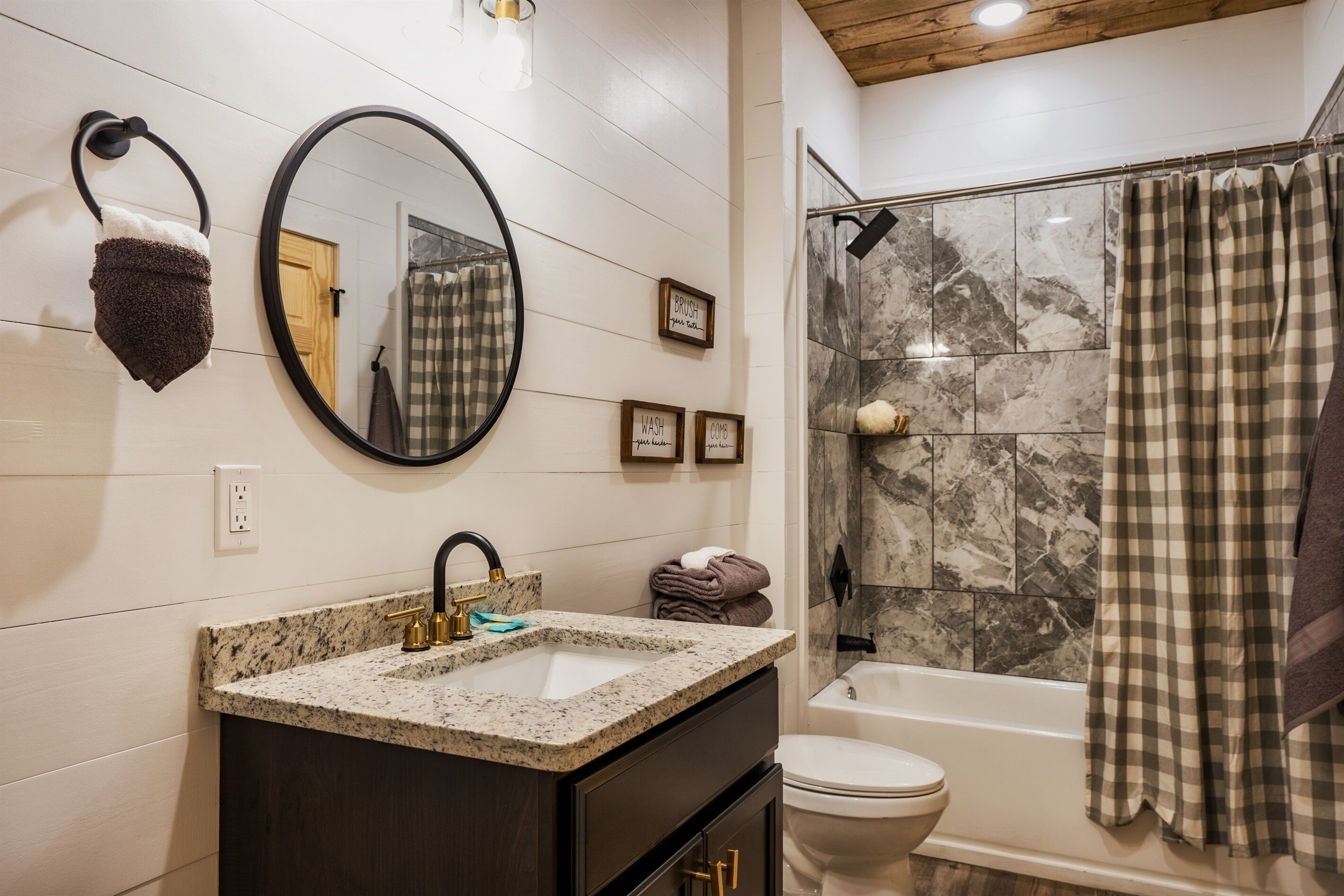 A bathroom with a granite sink, a black faucet, a mirror, a towel rack, a shower with a checkered curtain, and a toilet.