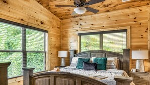 A wooden cabin bedroom with a bed, two windows, a ceiling fan, and a nightstand.