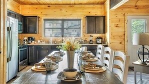 A wooden cabin interior with a dining table set for four, kitchen cabinets, and a window with blinds.