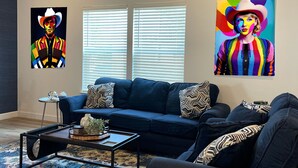 A modern living room with a dark blue sofa, a coffee table, and a patterned rug. There are two windows with blinds, a ceiling fan, and two vibrant paintings on the walls. - New Lovely Home in Lockhart with a Lounge (Lockhart)