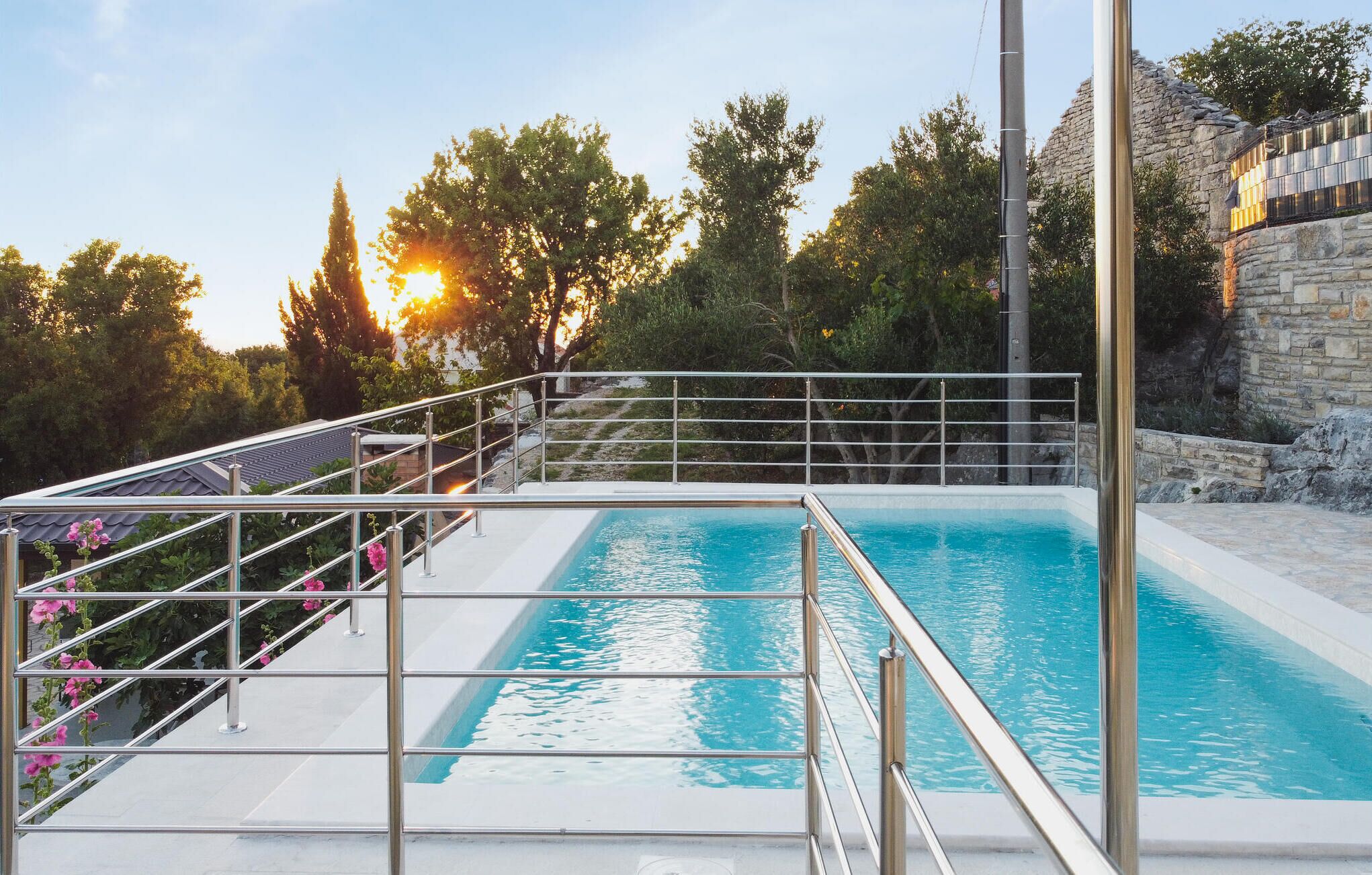A swimming pool with a metal railing, surrounded by greenery and a stone wall.