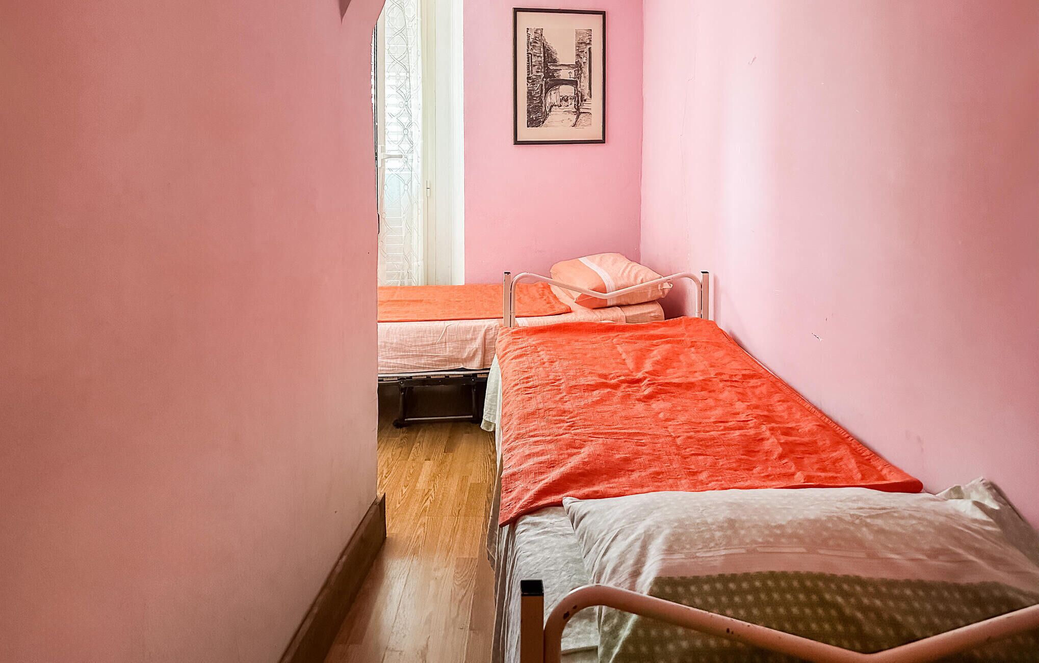 A narrow room with two single beds, one with an orange blanket and the other with a white blanket. There is a framed picture on the wall and a window with a metal grate.