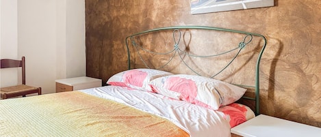 Cama bem arrumada com edredom floral e duas almofadas, mesa de cabeceira de madeira com gaveta, cabeceira de metal e uma pintura de paisagem urbana na parede.