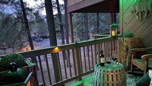 Una terraza de madera con barandilla, una mesa redonda con una botella y un vaso, un farol y un banco.