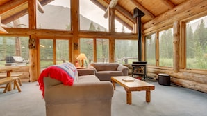 Ein Blockhaus-Interieur mit Sofa, Sessel und Tisch, großen Fenstern und einem Kaminofen.