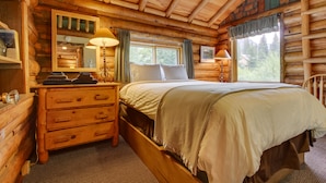 Ein Schlafzimmer in einer Blockhütte mit einem hölzernen Bett, einem Schminktisch, einem Spiegel und einem Fenster mit Vorhängen.