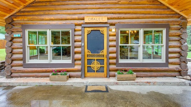 Eine Holzblockhütte mit einem Schild, auf dem 'CHALET' steht, zwei Fenstern an jeder Seite und hölzernen Pflanzgefäßen mit Blumen davor.