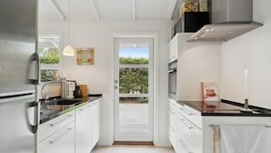 Ett smalt kök med vita skåp, ett kylskåp i rostfritt stål, en kaffebryggare och en utsikt över en trädgård genom en glasdörr.