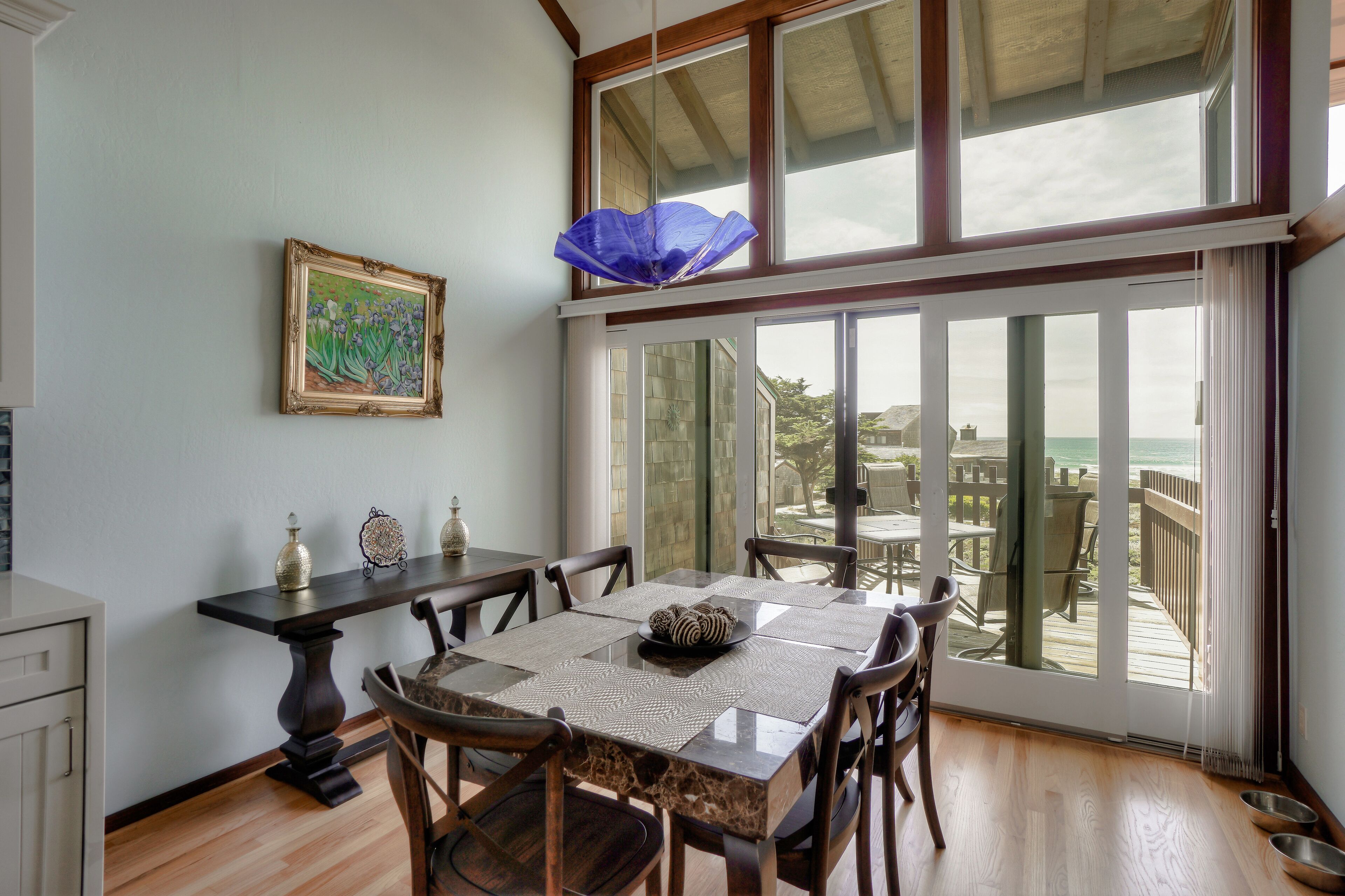 Un coin repas avec une table dressée pour quatre personnes, un tableau accroché au mur et une vue sur l’océan par de grandes fenêtres.