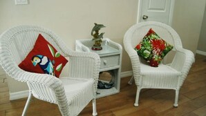 Deux fauteuils en osier blanc agrémentés de coussins colorés, une petite table d’appoint blanche ornée d’un objet décoratif et une porte fermée dans une pièce au plancher en bois.