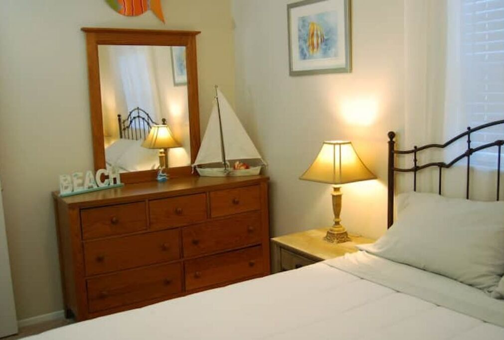 Une chambre avec une commode en bois, un lit recouvert de draps blancs, une table de chevet avec une lampe, un miroir et une maquette de voilier.