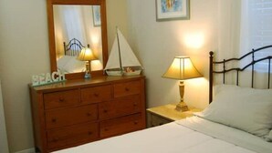 Une chambre avec une commode en bois, un lit recouvert de draps blancs, une table de chevet avec une lampe, un miroir et une maquette de voilier.