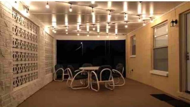 Un espace extérieur aménagé en patio, avec une table et des chaises, un mur en briques orné d’un motif en treillis et des guirlandes lumineuses suspendues au plafond.