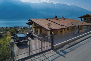 A residential building with a tiled roof, a fenced driveway, and a view of a lake and mountains in the background. - "Villa Imelda" spacious, bright apartments, garden or balcony, lake view (Limone sul Garda)