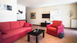Una sala de estar con un sofá rojo, un sillón rojo, una mesa de centro negra con una computadora portátil y obras de arte en las paredes.