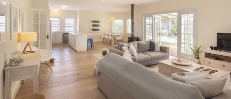 Una amplia sala de estar con un sofá grande color gris, una mesa de centro y un televisor. Se observa un área de cocina con gabinetes blancos y una mesa de comedor.