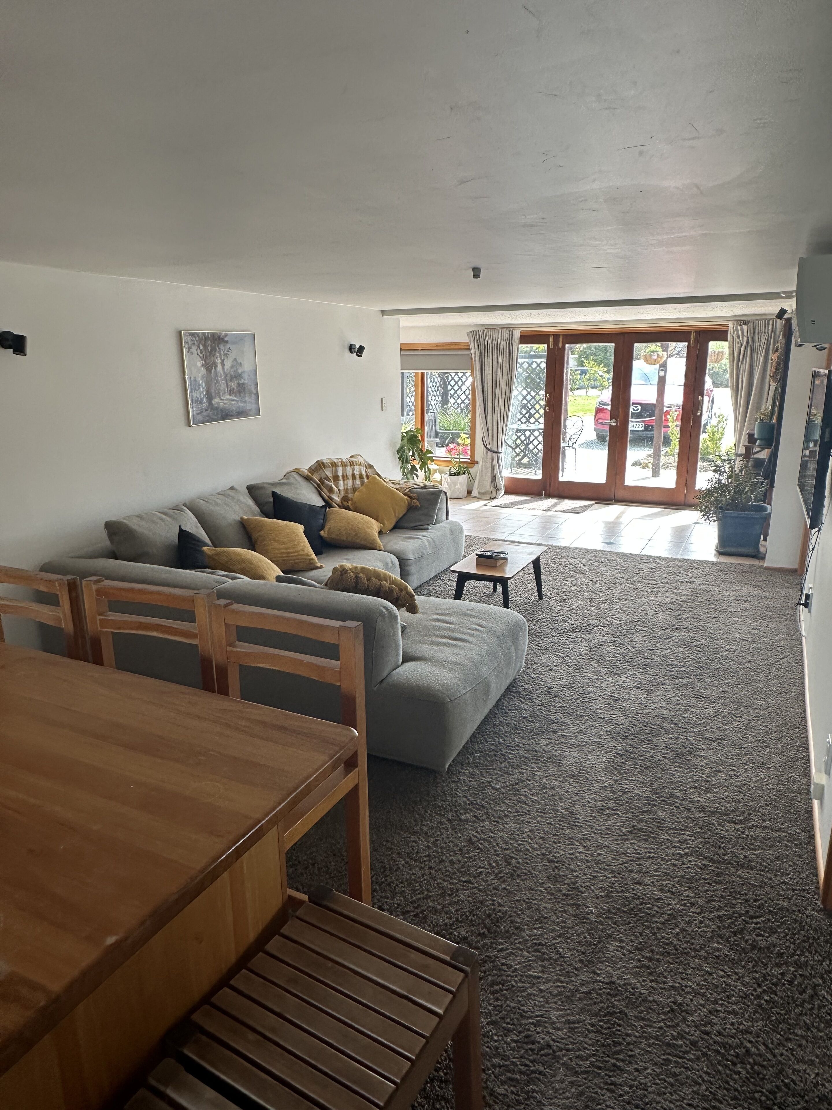 A living room with a sofa, a dining table, and chairs. There is a sliding glass door leading to a patio with a car parked outside.