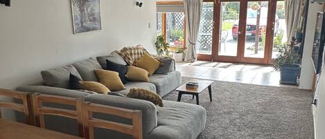 A living room with a sofa, a dining table, and chairs. There is a sliding glass door leading to a patio with a car parked outside.