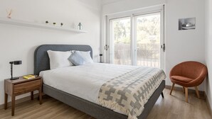 A modern bedroom with a bed, bedside table, chair, and a window with a view. - JOIVY Striking flat in Benfica (Lisboa)