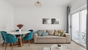A modern living room with a round dining table, teal chairs, a beige sofa with colorful cushions, and a large window with curtains. - JOIVY Striking flat in Benfica (Lisboa)